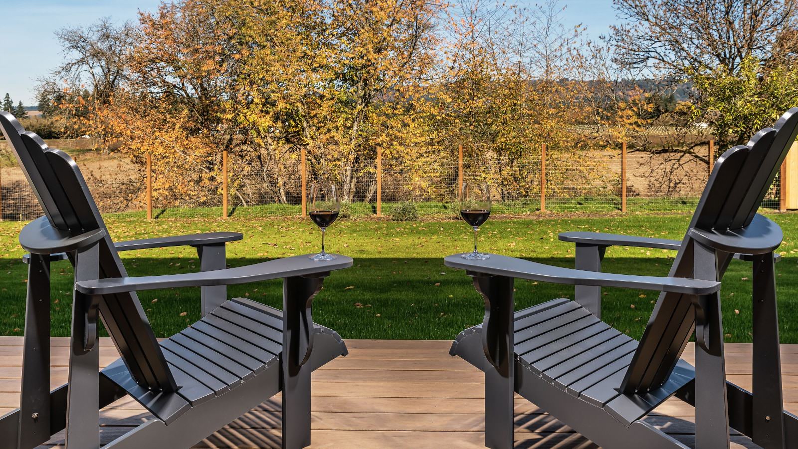 Two gray adirondack chairs each with a glass of red wine with bushes and trees in the background with fall color