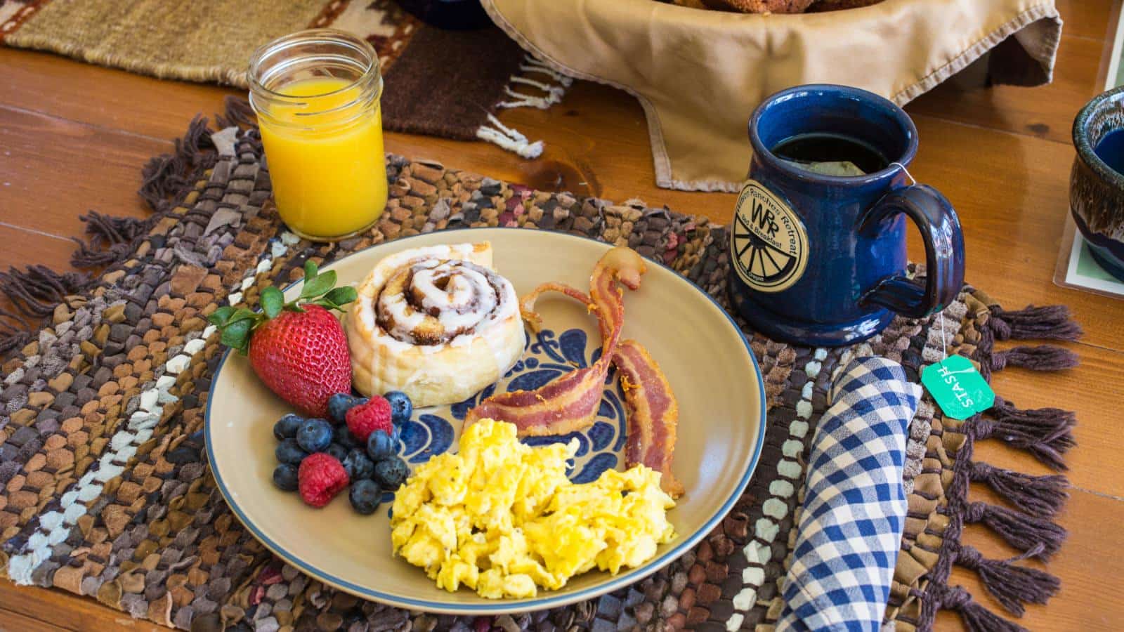 Close up view of light tan plate with scrambled eggs, bacon, cinnamon roll, and fruit