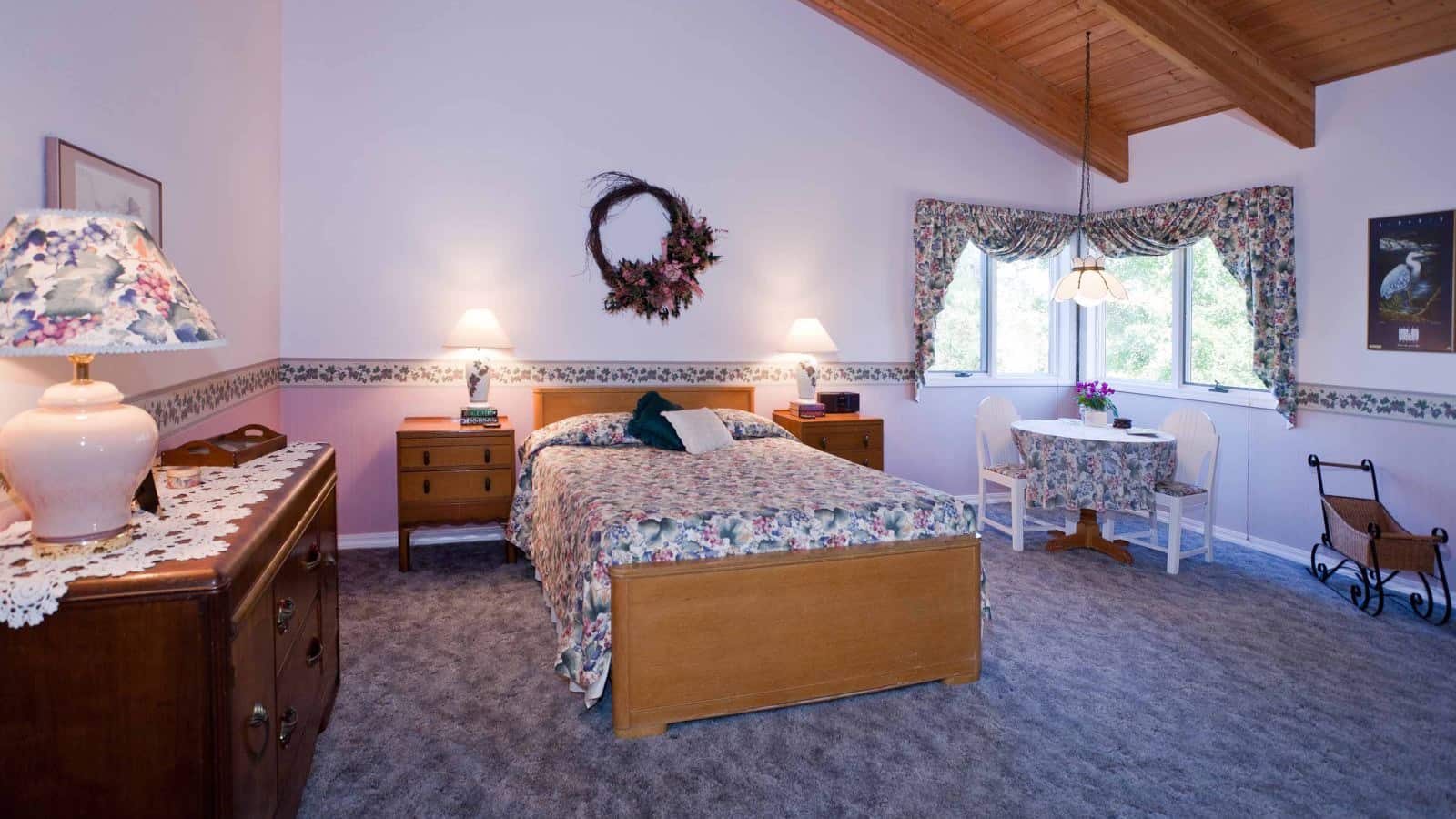 Bedroom with light colored walls, carpeting, light wooden bed, multicolored floral wedding, antique wooden dresser, and sitting area