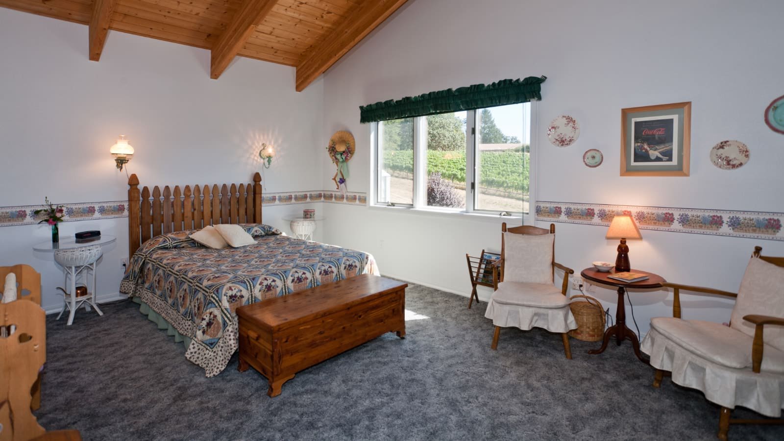 Bedroom with white walls, carpeting, wooden picket fence headboard, multicolored bedding, wooden trunk, and sitting area