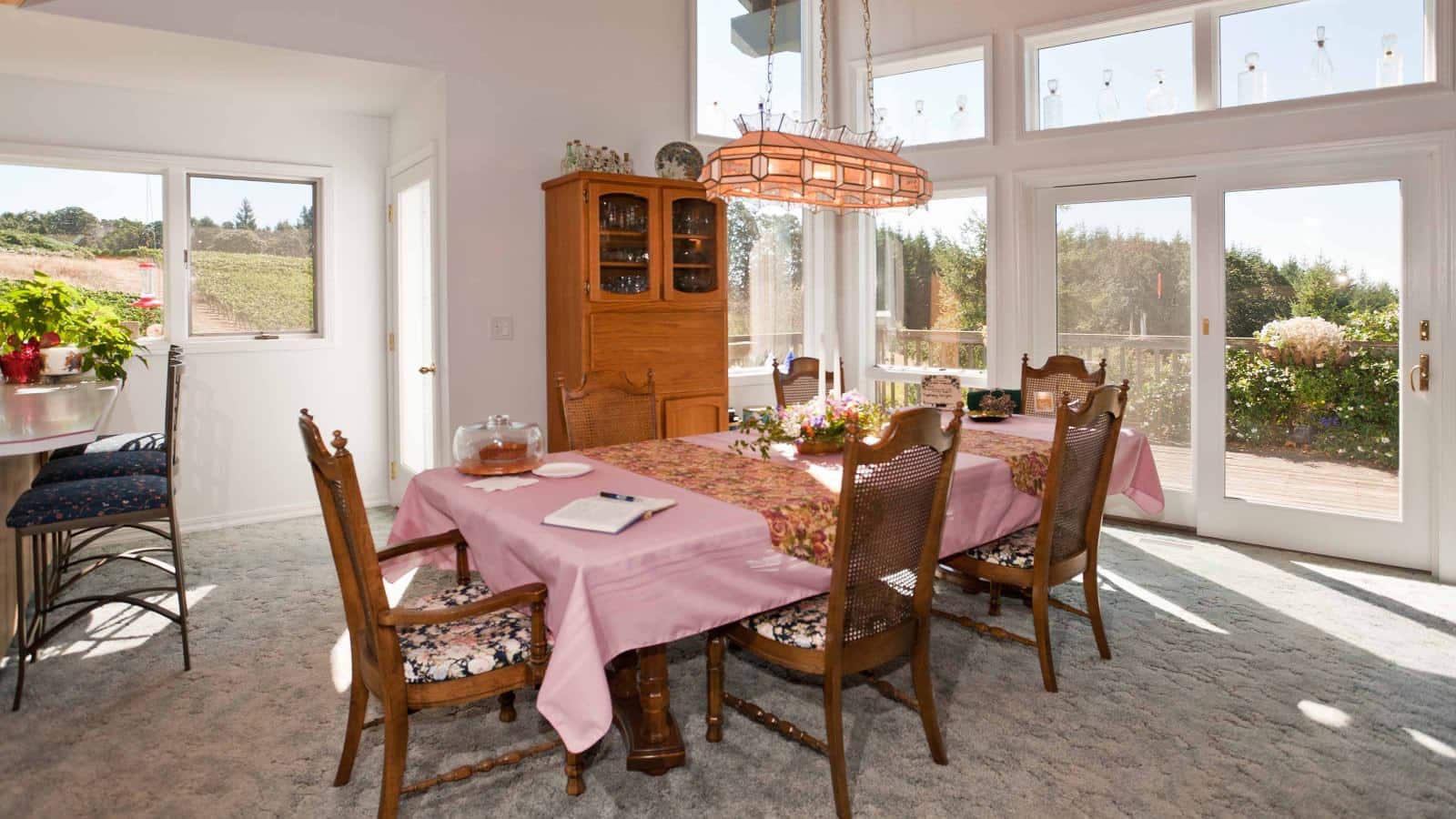 Dining room with white walls, carpeting, wooden table with pink tablecloth, wooden chairs, wooden china hutch, and many windows with views of the outside