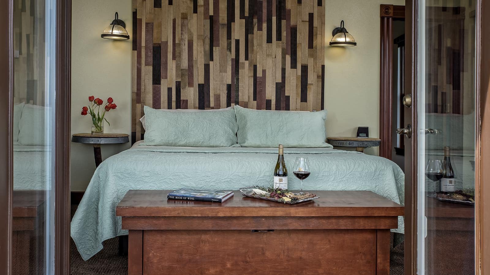 Bedroom with light colored walls, dark carpeting, wood accent wall behind bed, sea foam colored bedding, and large wooden trunk at foot of bed