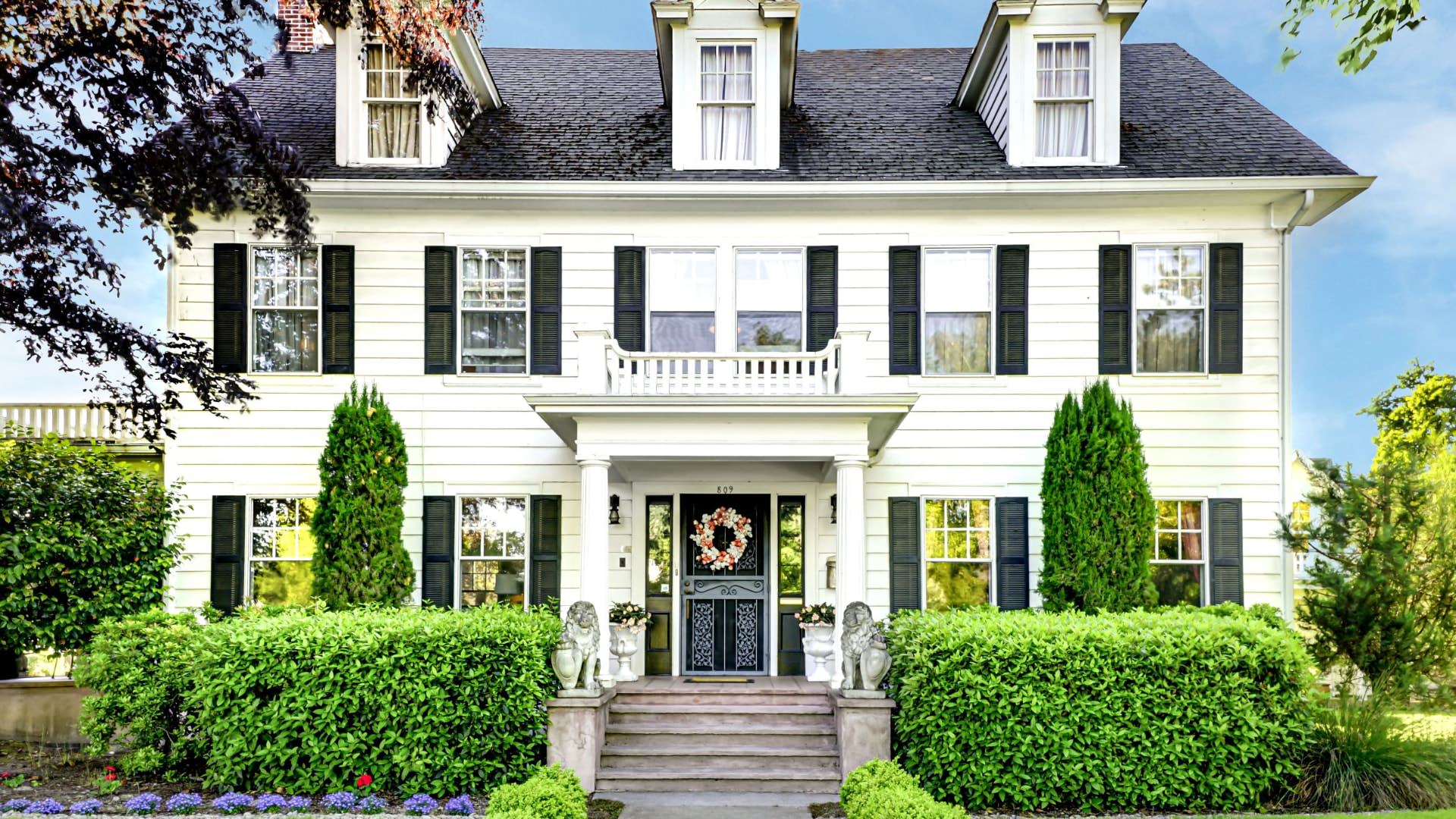 Exterior view of property painted white with dark shutters, steps up to entry, and surrounded by green shrubs and trees