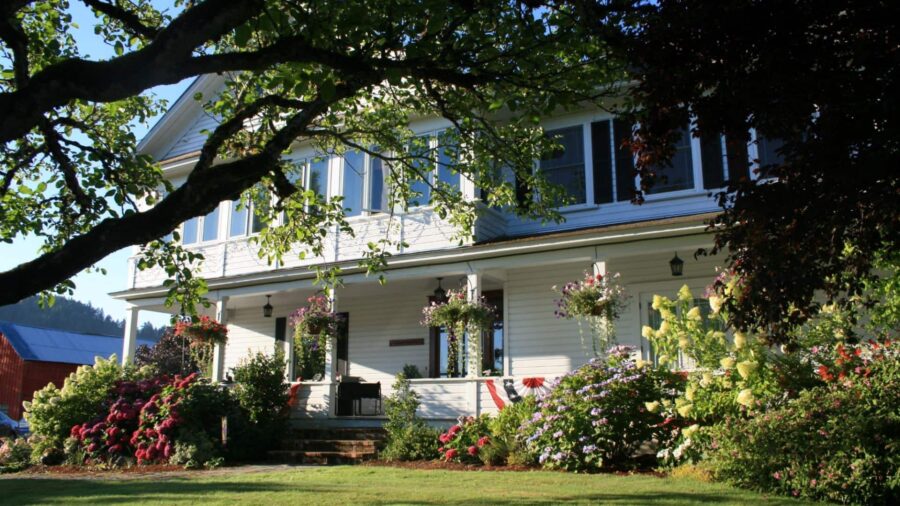 Exterior view of property painted white with white trim and dark shutters surrounded by lush bushes, flowers, and trees