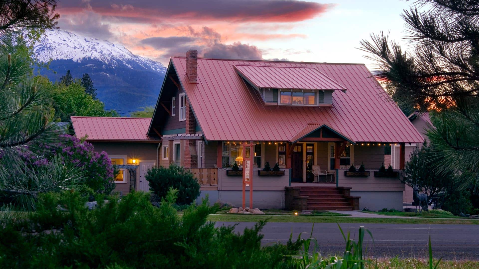 Exterior view of property surrounded by green grass, shrubs, and trees with snow-capped mountain in the background at dawn