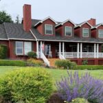 Exterior view of property painted red with white trim and green shutters surrounded by green grass, shrubs, flowers and trees