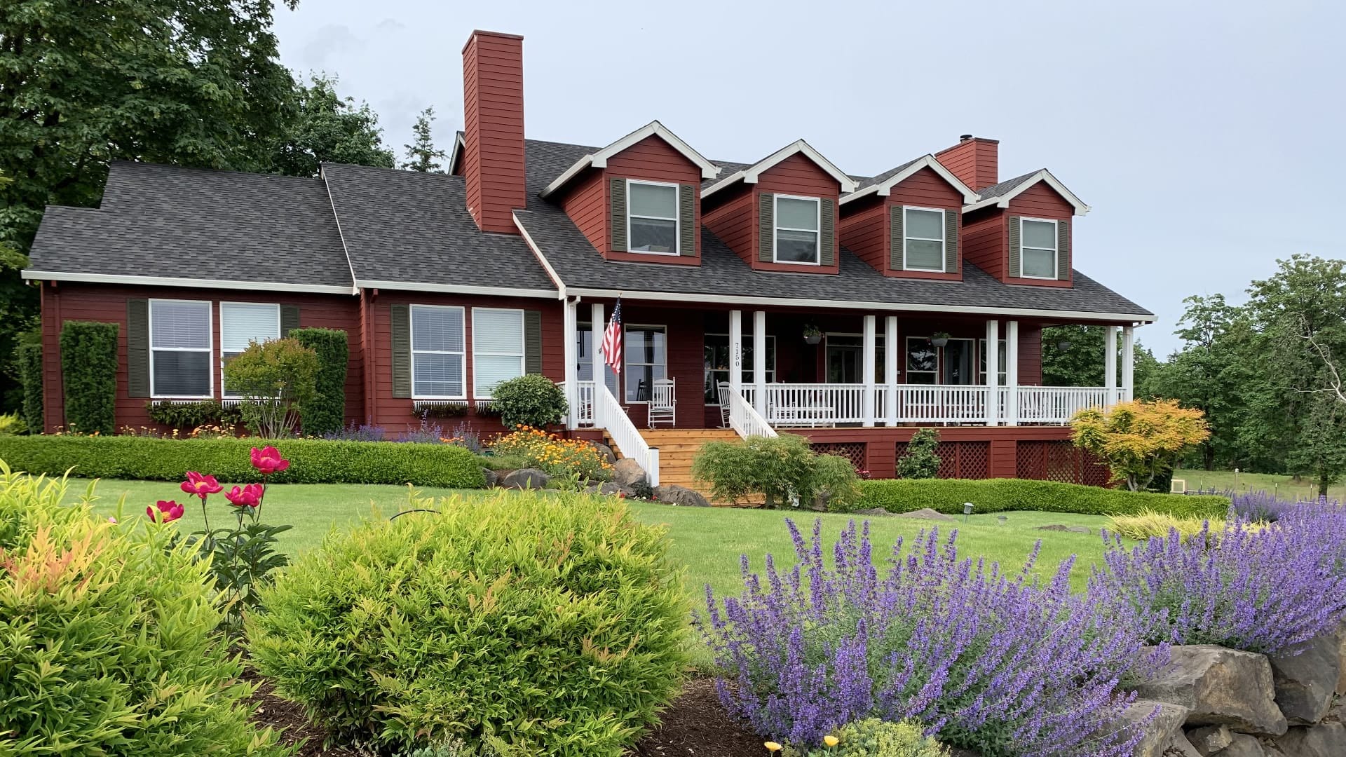 Exterior view of property painted red with white trim and green shutters surrounded by green grass, shrubs, flowers and trees