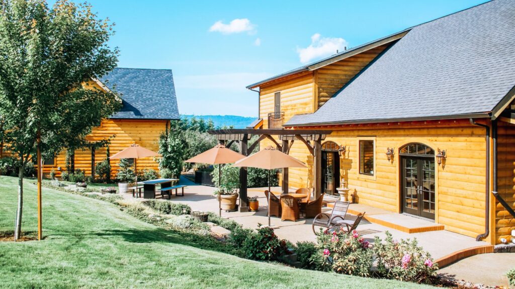 Exterior view of property with wooden log cabin siding, patio with pergola, patio furniture, and umbrellas surrounded by green grass, flowers, and trees