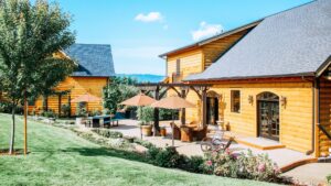 Exterior view of property with wooden log cabin siding, patio with pergola, patio furniture, and umbrellas surrounded by green grass, flowers, and trees