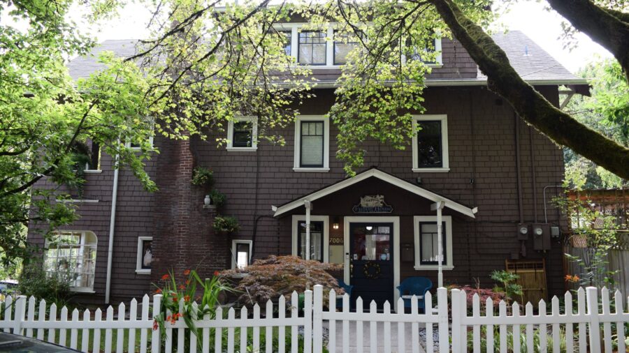 Exterior view of the property painted dark gray with white trim surrounded by green trees, shrubs, and white picket fence