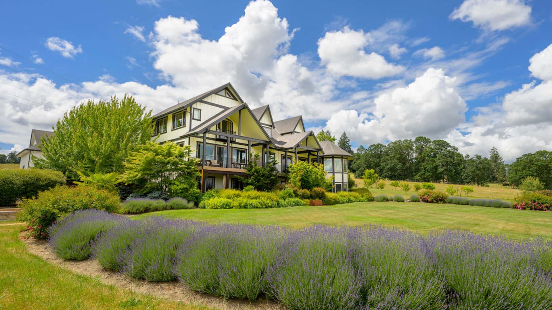 Exterior view of property painted light yellow with dark trim surrounded by large green lawn and many shrubs, trees, bushes, and flowers