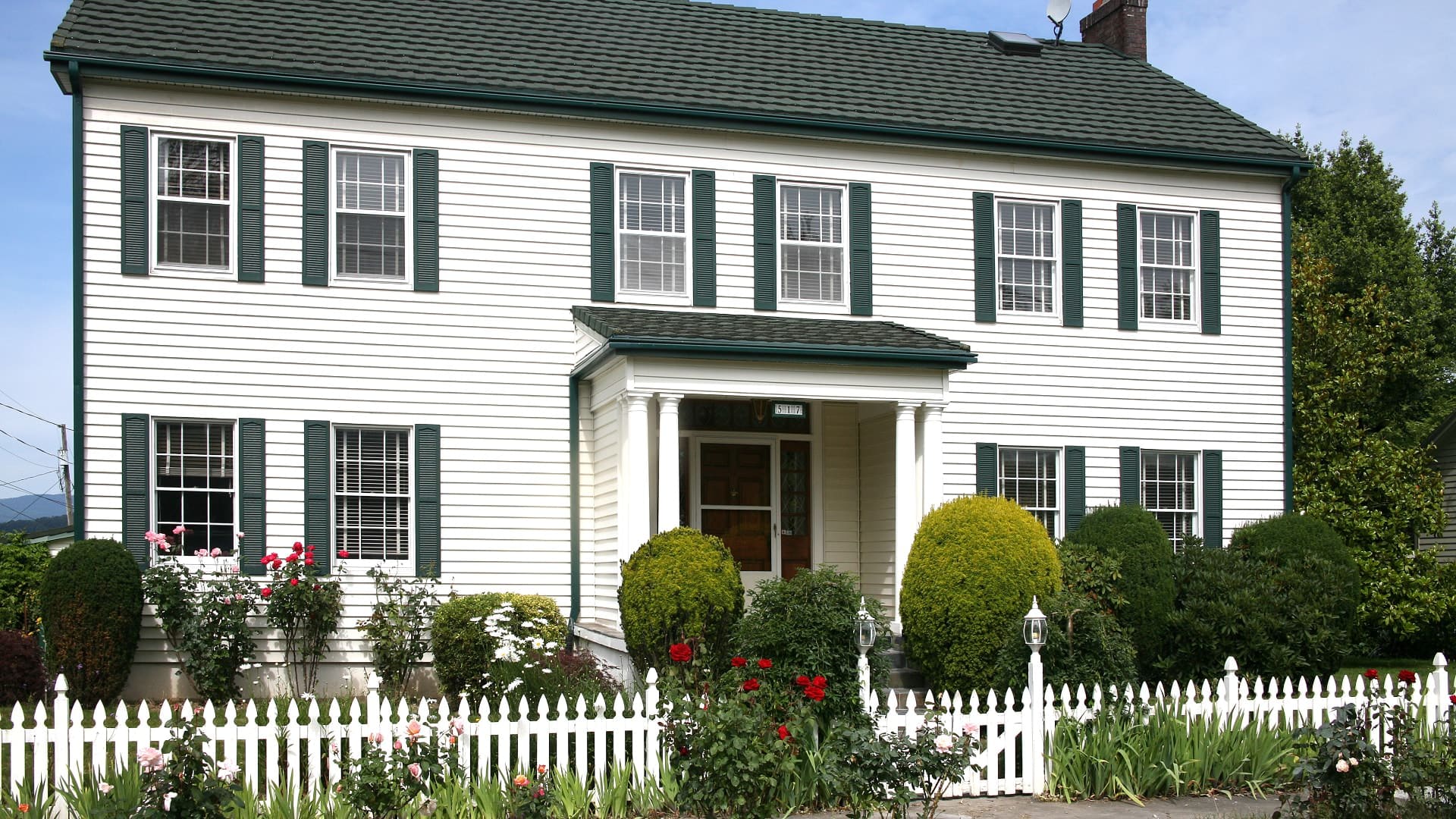 Exterior view of property painted white with green trim and shutters surrounded by green shrubs, bushes, flowers, and white picket fence