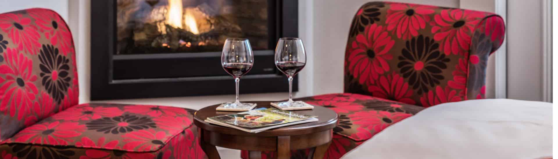 Close up view of two glasses of red wine on a small wooden table near two upholstered chairs with a red and black flower print and fireplace in the background