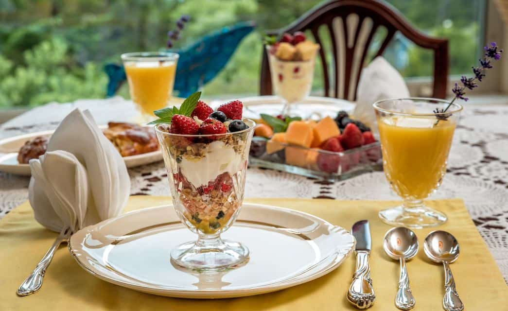 Close up view of fruit parfait in clear glass cup sitting on white plate with other breakfast dishes in the background
