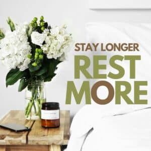 Stay longer rest more Close-up of a bedside table with a glass jar of white hydrangeas and greenery, an amber candle labeled “Sunday Campfire,” a smartphone, and crisp white bedding with a knit throw.