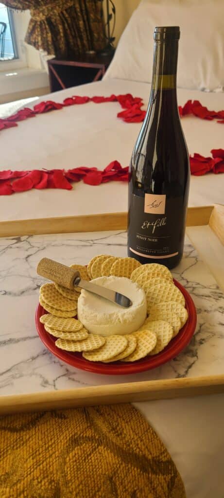 Truffle crackers, cheese and wine offered on white bedspread with red heart petals